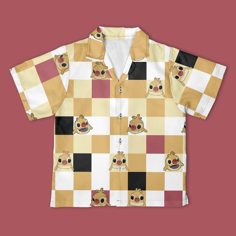 May include: A short-sleeved button-down shirt with a checkerboard pattern in shades of yellow, white, black, and maroon. The shirt features a repeating cartoon character design. The shirt has a collar and is buttoned up.