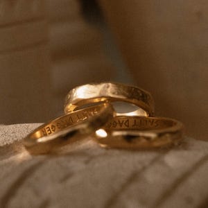 May include: Three gold-toned rings with a textured, hammered surface. The interior of the bands features the engraved text SALTY DAGGER. The rings are arranged in a cluster, showcasing their metallic finish and the visible inscription inside.