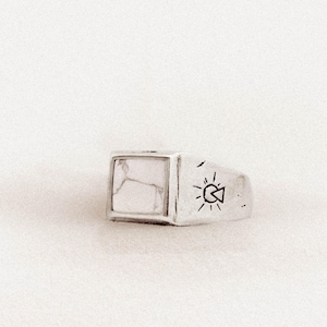 May include: A silver ring with a square white stone setting. The ring has a rectangular shape with a fish and sun design etched on the side. The white stone has a marbled appearance.