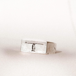 Könnte beinhalten: Ein silberner Ring mit rechteckigem Band. Der Ring hat ein kleines, geätztes Design eines Pinguins, der auf einem Surfbrett balanciert. Der Ring hat ein leichtes Vintage-Aussehen.