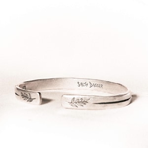 May include: A silver cuff bracelet with etched leaf designs and the text "SALTY DAGGER". The open-ended bracelet has a minimalist design and is likely made of sterling silver. The bracelet is a jewelry item.