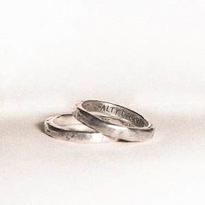 May include: Two silver rings with a textured finish are stacked on a white surface. The rings have the text "SALTY DAGGER" engraved on the outer band. The rings are likely for a couple or for everyday wear.