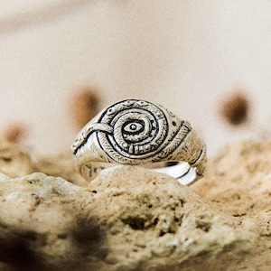 May include: A silver-toned metal ring featuring a textured, coiled snake design that forms a circular pattern. The centre of the spiral displays a small, distinct eye motif. The surface of the ring has an oxidised finish with dark recessed details.
