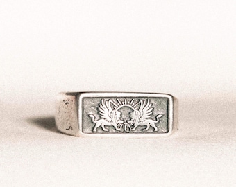 May include: A silver-toned rectangular ring featuring a detailed relief of two winged lions facing each other, with a sun-like emblem between them. The ring has a slightly worn appearance, suggesting an antique or vintage style.
