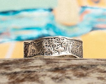 May include: A silver-colored ring with a textured surface and intricate embossed designs. The ring is set against a blurred background of blue, yellow, and white colors. The ring appears to be made of metal.
