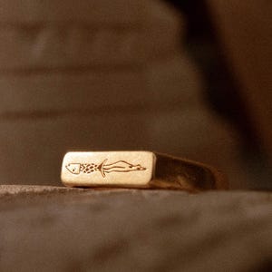 Könnte beinhalten: Ein goldfarbener, rechteckiger Ring mit einem gravierten Fischmuster. Der Ring hat eine glatte, polierte Oberfläche und eine leicht abgerundete Form. Das Fischmuster befindet sich mittig auf der Oberseite des Rings.