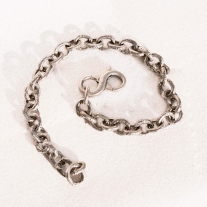 May include: A silver chain bracelet with a textured, aged look. The bracelet has a series of interconnected oval links and a secure clasp. The chain is displayed on a white surface.