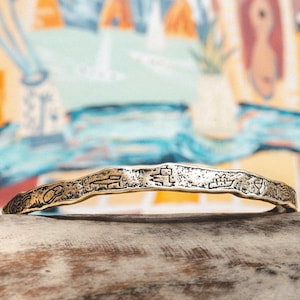 Puede incluir: Una pulsera de metal dorado con una superficie irregular y texturizada. La pulsera está adornada con pequeños símbolos grabados y descansa sobre una superficie de madera. El fondo presenta una pintura abstracta colorida.