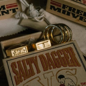 May include: Gold-toned rings and rectangular rings with embossed designs are displayed in an open box. The box has the words "SALTY DAGGER" printed on it. Other boxes and drawstring bags are in the background.
