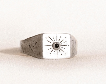 May include: A silver signet ring with a square face. The face features a sunburst design with a central black dot. The ring has a slightly distressed, vintage appearance.