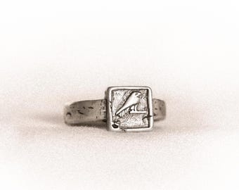 May include: A silver-toned ring featuring a square-shaped face with an engraved bird design. The band has a textured, weathered finish, and the central square plaque displays a detailed depiction of a bird perched on a branch in profile.