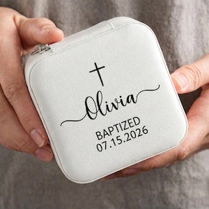 May include: A white, square jewelry case with a zipper closure. The case features a black cross and the name "Olivia" in script, along with "BAPTIZED 07.15.2026". The case is held in someone's hands.