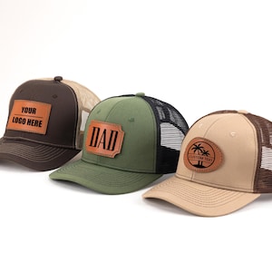Puede incluir: Tres gorras de camionero con parches de cuero. Una es marrón con "YOUR LOGO HERE", otra es verde con "DAD", y la última es beige con un diseño de palmera y "CUSTOM TEXT". Todas tienen la parte trasera de malla.