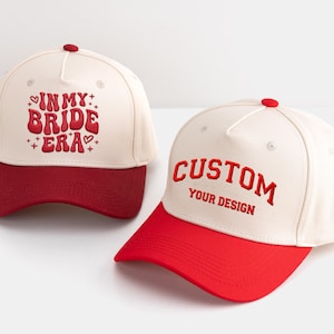 May include: Two baseball caps. One is cream-coloured with a red brim and the text "IN MY BRIDE ERA" in red. The other is cream-coloured with a red brim and the text "CUSTOM YOUR DESIGN" in red.