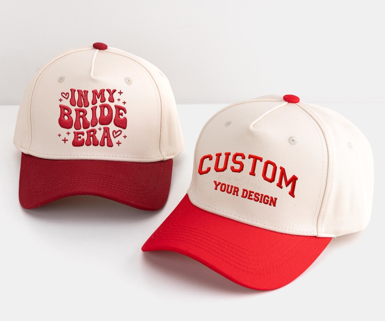 May include: Two baseball caps. One is cream-coloured with a red brim and the text "IN MY BRIDE ERA" in red. The other is cream-coloured with a red brim and the text "CUSTOM YOUR DESIGN" in red.