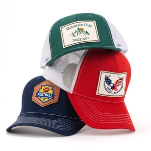 Op de afbeelding: Drie baseballpetten op elkaar gestapeld. De bovenste pet is groen met een witte mesh achterkant en een patch met de tekst "Mountain Club SINCE 2023". De middelste pet is rood met een witte mesh achterkant en een adelaars patch. De onderste pet is marineblauw met een voetbal patch.