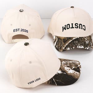 Op de afbeelding: Drie crèmekleurige baseballpetten met camouflagekleurige kleppen. Eén pet heeft de tekst CUSTOM in het zwart, een andere toont EST. 2026 en de derde bevat de tekst YOUR LOGO. Elke pet heeft een kleine camouflagekleurige knoop op de bovenkant.