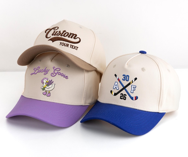 May include: Three baseball caps in cream, with different coloured brims. One cap has "Custom" embroidered on the front, another has "Lucky Goose" with a goose graphic, and the third has crossed hockey sticks and numbers.