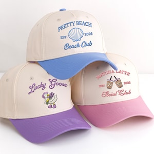 May include: Three baseball caps with cream-coloured crowns and coloured brims. One has a blue brim and the text "Pretty Beach Beach Club". Another has a purple brim and the text "Lucky Goose". The third has a pink brim and the text "Matcha Latte Social Club".