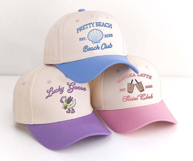 May include: Three baseball caps with cream-coloured crowns and coloured brims. One has a blue brim and the text "Pretty Beach Beach Club". Another has a purple brim and the text "Lucky Goose". The third has a pink brim and the text "Matcha Latte Social Club".