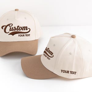 May include: Two beige baseball caps with brown visors. The word "Custom" is embroidered in brown script on the front of each cap, with "YOUR TEXT" below. The caps have a curved brim and a button on top.