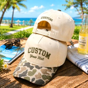 Peut inclure: Deux casquettes de baseball couleur crème avec des visières camouflage. La casquette du bas porte le texte brodé "CUSTOM Your Hat" en vert foncé, tandis que celle du haut affiche "CUSTOM" à l'arrière. Les deux ont des fermetures réglables.