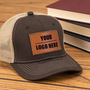 May include: A brown trucker hat with a beige mesh back and a brown leather patch. The patch has the text "YOUR LOGO HERE" in black. The hat is on a wooden surface next to a stack of books.