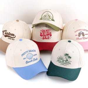 May include: Several baseball caps in shades of beige, red, pink, blue, and green. Each cap has embroidered text and designs, including "Custom," "Pretty Beach," and "Matcha Latte." The caps are displayed on a white background.