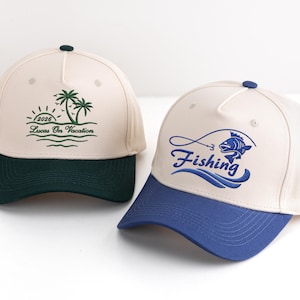 May include: Two baseball caps with contrasting brims. One is cream-coloured with a dark green brim and a palm tree design with the text "2026 Lucas On Vacation". The other is cream-coloured with a blue brim and the word "Fishing" with a fish graphic.