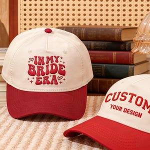 May include: Two baseball caps with a white crown and a red brim. One cap has the embroidered text "IN MY BRIDE ERA" in red. The other cap has the embroidered text "CUSTOM YOUR DESIGN" in red.