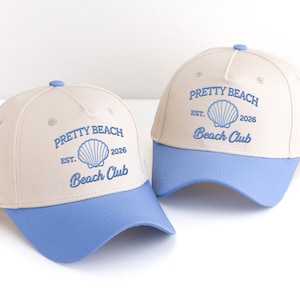 May include: Two baseball caps with a cream-coloured crown and a light blue brim. The caps feature the text "PRETTY BEACH Beach Club" with a seashell graphic. A small blue button is on top of each cap.