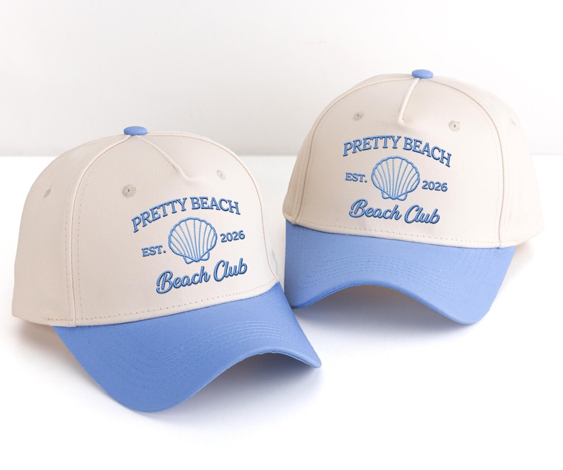 May include: Two baseball caps with a cream-coloured crown and a light blue brim. The caps feature the text "PRETTY BEACH Beach Club" with a seashell graphic. A small blue button is on top of each cap.