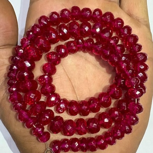 May include: A necklace of faceted, deep red beads. The beads are strung together, forming multiple strands. The necklace is displayed on a hand, showcasing the color and texture of the gemstones. The beads have a sparkling, polished appearance.