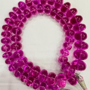 May include: A strand of translucent, oval-shaped beads in a vibrant magenta hue. The beads are strung together, forming a necklace or bracelet. The ends of the strand are finished with silver-colored clasps and pink string.