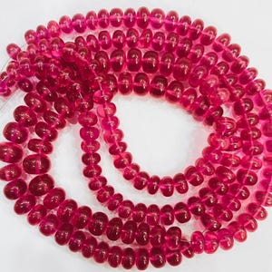 May include: Strands of translucent, deep pink beads. The beads are oval-shaped and strung together, creating a layered effect. The beads have a glossy finish and are arranged in a circular pattern. The overall theme is jewelry.