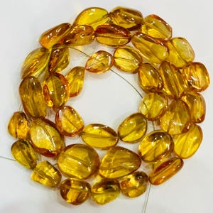 May include: A necklace of polished amber beads. The beads are various shapes and sizes, ranging from oval to irregular, and are strung together on a thin, nearly invisible cord. The amber has a warm, golden-yellow color with variations in transparency.