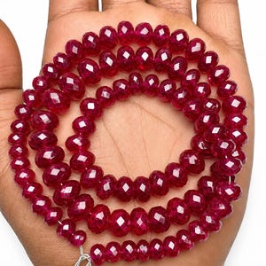Peut inclure: Un collier de perles circulaires facettées d'un rouge profond disposées en spirale. Les perles présentent une qualité translucide et une surface réfléchissante, révélant de multiples petites facettes géométriques qui captent la lumière.