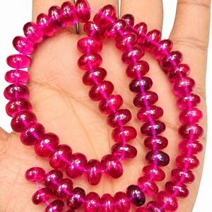 May include: A strand of faceted, translucent, deep pink gemstone beads arranged in a circular pattern. Each bead features a rounded, disc-like shape with a polished surface that reflects light, strung together on a thin, bright pink thread.