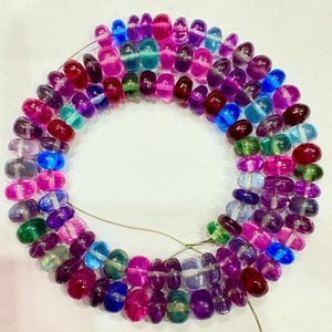 May include: A strand of colorful gemstone beads in shades of purple, pink, blue, and green. The beads are oval-shaped and strung together, creating a circular arrangement. The beads are translucent, allowing light to pass through.