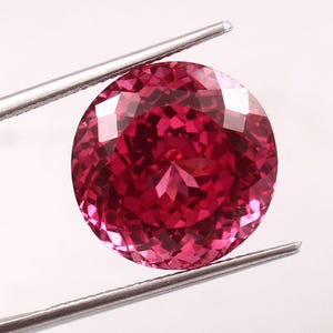 Może przedstawiać: Oszlifowany, okrągły, głęboko różowy kamień szlachetny trzymany jest przez srebrną pęsetę. Szlif kamienia szlachetnego tworzy wiele odbijających światło powierzchni, nadając mu wspaniały wygląd. Klejnot ma żywy odcień czerwono-różowy.