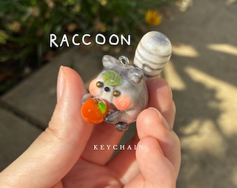 Mapache - dije de arcilla polimérica hecho a mano, lindo llavero de animal esponjoso sosteniendo una fruta, mapache gris regordete, escultura de arcilla original y caprichosa