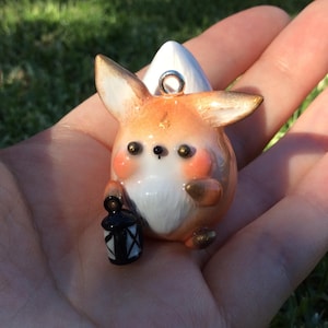 Puede incluir: Figura de zorro artesanal con detalles en naranja, blanco y dorado, sosteniendo una linterna negra. El zorro tiene orejas grandes y mejillas rosadas. La figura es un colgante con un lazo plateado para colgar.