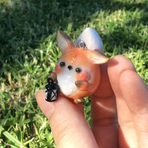 Puede incluir: Un colgante de zorro artesanal con detalles en naranja, blanco y dorado, sosteniendo una peque&ntilde;a linterna negra. El zorro tiene ojos grandes y oscuros y una cola blanca y esponjosa. Es una figura en miniatura.