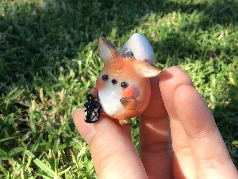 Puede incluir: Un colgante de zorro artesanal con detalles en naranja, blanco y dorado, sosteniendo una peque&ntilde;a linterna negra. El zorro tiene ojos grandes y oscuros y una cola blanca y esponjosa. Es una figura en miniatura.