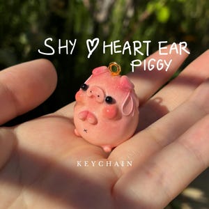 May include: A pink piggy keychain with heart-shaped ears, black eyes, and a gold-coloured loop. The text "SHY HEART EAR PIGGY KEYCHAIN" is written in white cursive above and below the pig figurine.