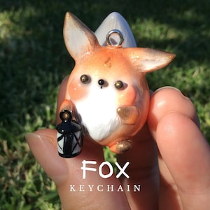 Puede incluir: Un llavero de zorro artesanal con detalles en naranja, blanco y dorado. El zorro sostiene una linterna en blanco y negro. El texto "FOX KEYCHAIN" se muestra debajo.