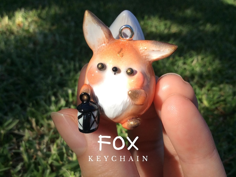 Puede incluir: Un llavero de zorro artesanal con detalles en naranja, blanco y dorado. El zorro sostiene una linterna en blanco y negro. El texto "FOX KEYCHAIN" se muestra debajo.