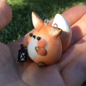 Puede incluir: Figura de zorro artesanal con detalles en naranja, blanco y dorado, sosteniendo una peque&ntilde;a linterna negra. El zorro tiene una cola blanca y est&aacute; dise&ntilde;ado con un estilo lindo y de dibujos animados. Tiene un peque&ntilde;o lazo plateado para colgar.