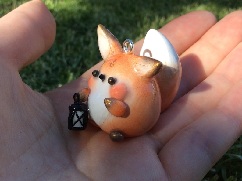 Puede incluir: Figura de zorro artesanal con detalles en naranja, blanco y dorado, sosteniendo una peque&ntilde;a linterna negra. El zorro tiene una cola blanca y est&aacute; dise&ntilde;ado con un estilo lindo y de dibujos animados. Tiene un peque&ntilde;o lazo plateado para colgar.