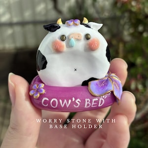 May include: A white and black cow figurine with small horns and pink cheeks, resting in a purple circular base holder. The base features a purple flower and the text "COW'S BED" in white lettering. The cow has a small blue teardrop detail.